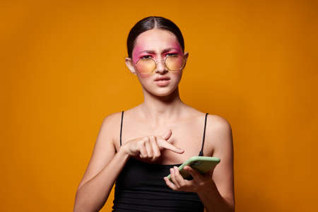 fashionable woman with phone pink face makeup posing attractive look glasses yellow background...