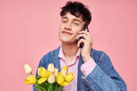 young guy in a denim jacket with a bouquet of flowers talking on the phone pink background