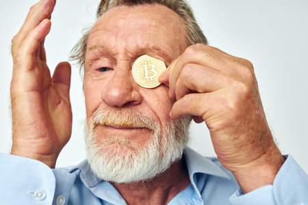 Senior grey-haired man finance gold coins bitcoin near face light backgroundの写真素材