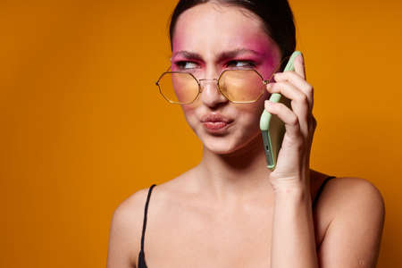 fashionable woman with phone pink face makeup posing attractive look glasses yellow background unalteredの写真素材