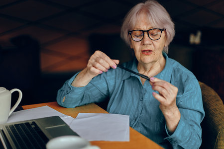 Portrait of an elderly woman with glasses sits at a table in front of a laptop Freelancer works unalteredの写真素材