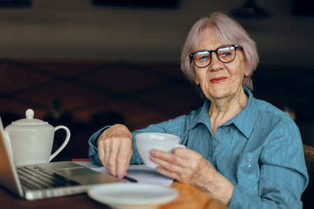 Portrait of an elderly woman with glasses sits at a table in front of a laptop Freelancer works unalteredの写真素材