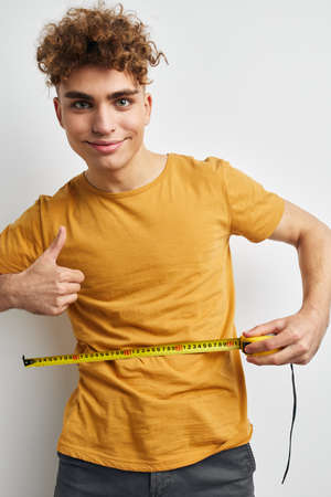Attractive man measuring tape measure in yellow t-shirt light backgroundの写真素材