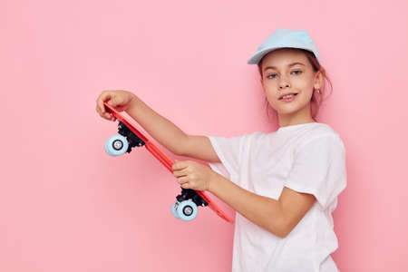 Portrait of happy smiling child girl cap in white t-shirt skateboard pink background