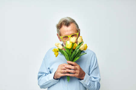 Senior grey-haired man in a blue shirt with a bouquet of flowers light background