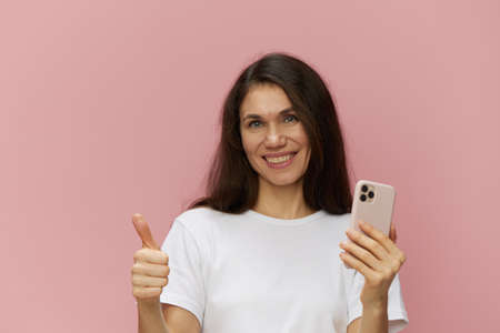 beautiful cute woman with a trendy phone in her hands stands on a pink background in a white...