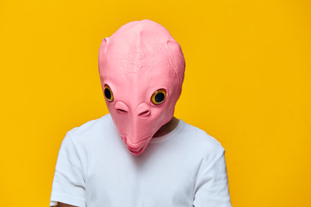 a man in a funny rubber mask looks at the floor while standing on a yellow backgroundの写真素材