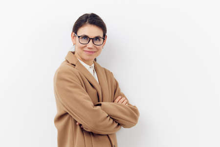 a beautiful, happy, brunette woman in glasses for vision stands in a brown stylish coat and smiles cutely looking at the camera with her arms folded on her chest.の写真素材