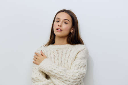 horizontal photo of a beautiful, gentle young woman, brunette standing in a white, warm, stylish sweater on a light background, with a hand on her shoulder, looking pleasantly into the cameraの写真素材