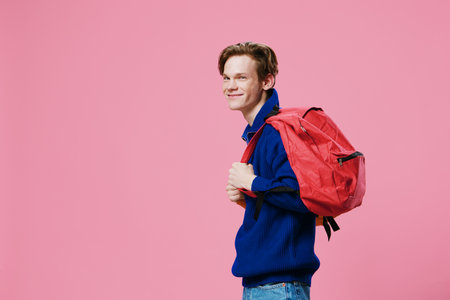 a happy guy in a blue sweater stands sideways to the camera with a red backpack on his back. Horizontal photo on a pink background with empty space for an insert for advertising textの写真素材