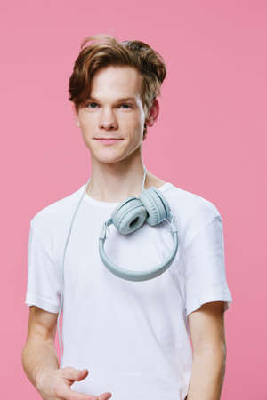 beautiful young man in a white T-shirt on a pink background with headphones around his neck smiling...