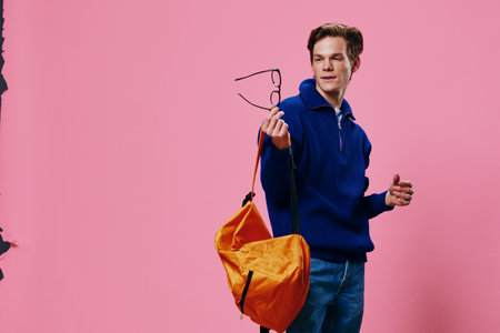 a handsome guy student with a bright backpack looks at his glasses with contempt while standing on a...