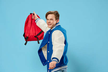 funny guy student in a trendy jacket holds his backpack on outstretched hand and smiles while...