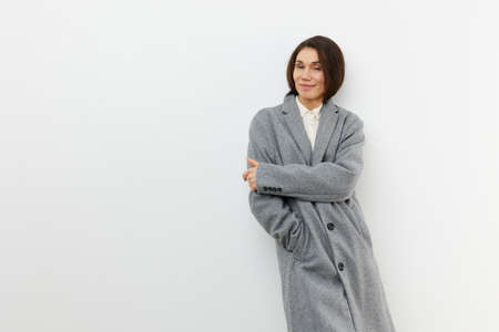 a horizontal portrait on a light background in a long gray coat stands smiling at the camera in a closed poseの写真素材
