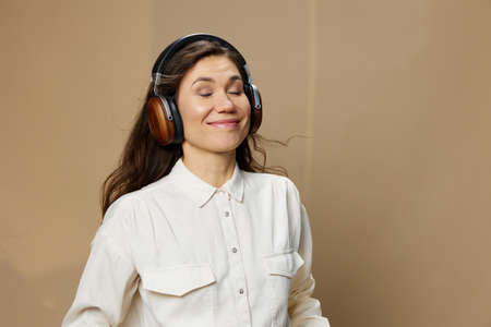 a happy, beautiful woman in a white shirt stands listening to music with headphones happily closing her eyes with pleasure while standing on a beige backgroundの写真素材
