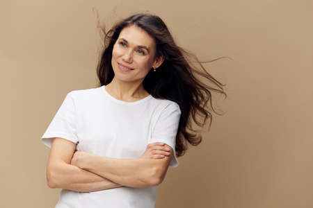 pleasant, funny woman with long well-groomed hair flying in the wind stands in a white T-shirt with her arms folded on her chest and smiles broadly with beautiful bright teeth with her eyes closedの写真素材