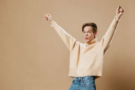 very happy enthusiastic young man in a long fluffy beige sweater raises his hands up expressing his emotionsの写真素材