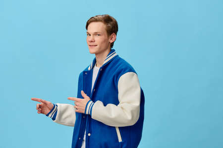 a young, handsome student boy in a blue bomber jacket points his fingers towards the space for text. Studio photo on a plain backgroundの写真素材