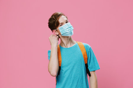 handsome guy in a blue t-shirt and with backpack on his back puts on a medical mask on his face while standing on a pink background. Horizontal portrait with empty space for advertising text insertionの写真素材