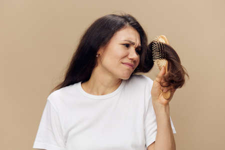A sad, upset woman stands with tangled hair in a comb and with an emotionally depressed face trying to untangle her hair standing on a beige backgroundの写真素材