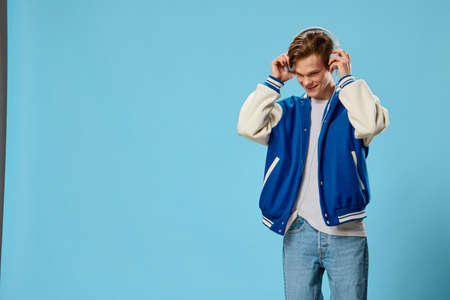 cute, handsome guy in a bomber jacket listens to music in big headphones holding them with his hands. Studio photography on a blue background with space for an advertising mockupの写真素材