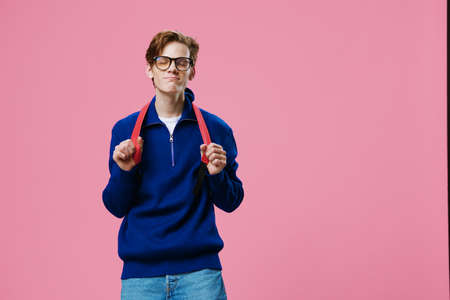 a student guy with a backpack on his back stands on a pink background in an autumn sweater and with...