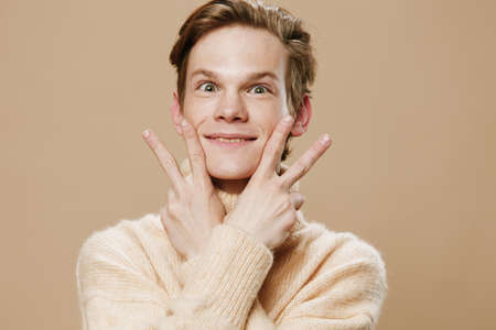 funny, funny guy in a beige sweater makes a funny face closing his eyes with his fingers. Horizontal photo on the theme of expressing emotionsの写真素材