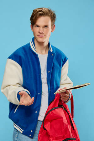 a pensive student in a blue stylish bomber jacket stands with a notebook in his hands and looks away. Vertical photograph on a light blue backgroundの写真素材