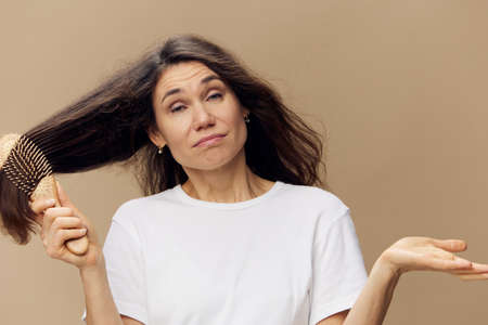 a sad, upset, emotional woman stands in a white cotton T-shirt and screams while combing her long hair. Horizontal photo on beige background with empty spaceの写真素材
