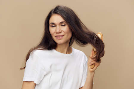 portrait of a beautiful woman in a white T-shirt on a beige background combing her long hair with a...