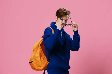 horizontal photo of a guy in a blue sweater, adjusting his glasses, with a backpack on his back. Studio photo on a uniform pink background with empty space for an advertising mockupの写真素材