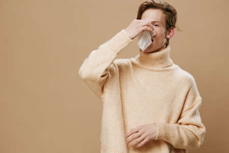 a guy in a beige sweater is sick and sneezes into a handkerchief. Horizontal photo with empty space for advertising mockupの写真素材