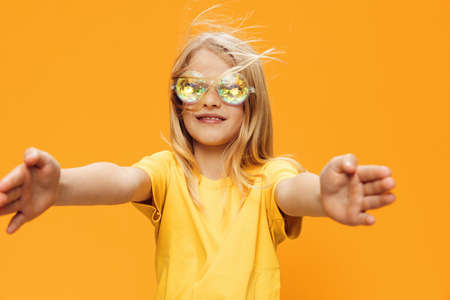 funny, playful girl posing standing on a bright background in yellow clothes and shiny, carnival glasses for a party and the wind blows her blond hair. Horizontal studio photographyの写真素材