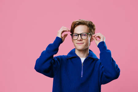 a young man with a beautiful hairstyle in a blue sweater adjusts his black-rimmed glasses while...