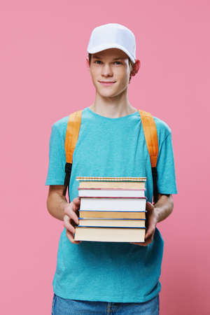 a handsome, funny guy in a blue t-shirt and a cap on his head stands holding textbooks in his hands with a backpack on his backの写真素材