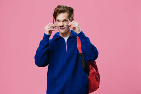 a joyful, handsome student guy in a blue jacket and with a red backpack on his shoulder puts on black glasses for vision. Pink background with space for text.の写真素材