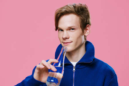 cute, handsome guy drinks water from a glass bottle. Studio photo on a plain pink backgroundの写真素材