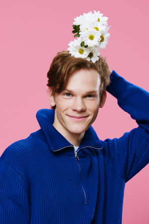 a funny guy in a blue sweater on a pink background holds a bouquet of large white daisies in his...