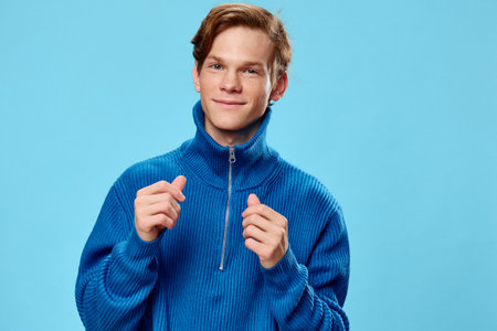 portrait of a handsome red-haired guy in a blue zip-up sweater standing against a light blue background and smiling at the cameraの写真素材