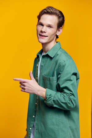 A male blond student in a T-shirt and a green shirt stands on a yellow background, smiles and looks...