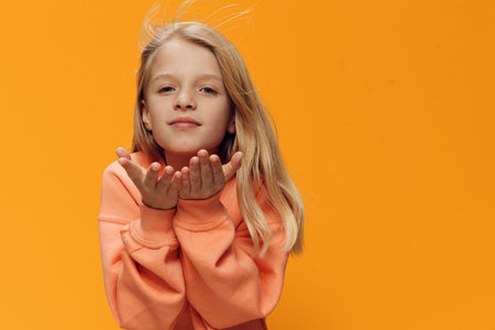 a beautiful, sweet, cheerful girl of school age, blonde, stands in an orange sweater on a yellow background sending an air kiss to the camera. Horizontal photo with empty space for textの写真素材