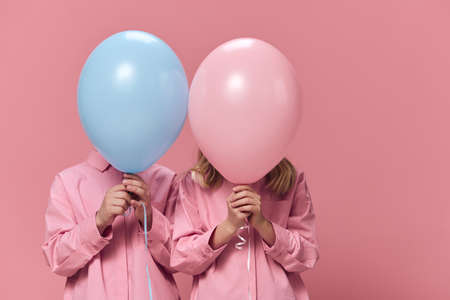 school-age children stand holding balloons in their hands, covering their faces with them....