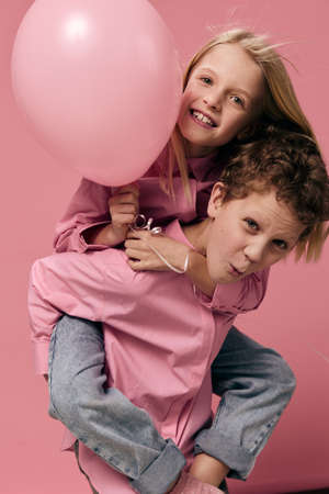 joyful, happy children of school age, on a pink background in pink clothes play and the boy rolls the girl with a balloon on her backの写真素材