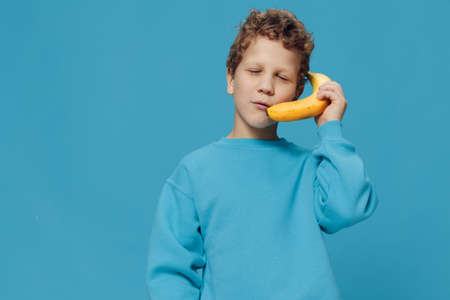 funny boy in blue clothes stands on a blue background and pretends to talk on a bananaの写真素材