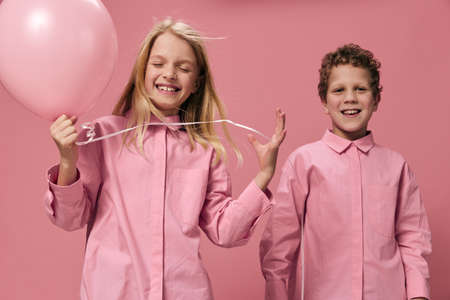 beautiful joyful children brother and sister stand on a pink background in pink clothes with a pink balloon in their hand and the girl closed her eyes from enjoymentの写真素材