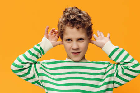 horizontal portrait of a cute, funny, playful, curly-haired boy of school age in a striped long-sleeved t-shirt on an orange background, smiling broadly, looking at the camera and grimacingの写真素材