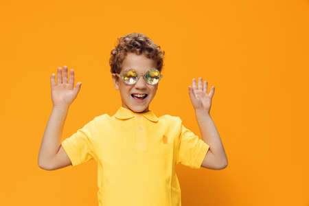school age boy is standing in a yellow polo in funny, shiny party glasses with a very surprised expression on his face and holding his head with his hands. Horizontal studio photo with empty spaceの写真素材