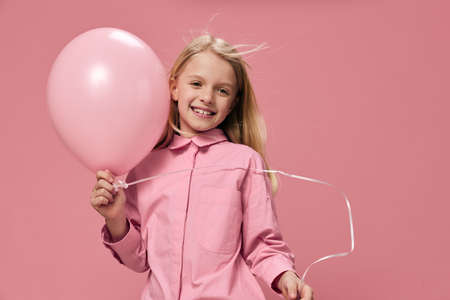 a happy, joyful school-age girl stands on a pink background with a big balloon in her hand and smiles broadly while looking at the camera.の写真素材