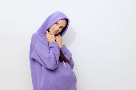 an interested woman stands on a white background in a purple tracksuit put a hood on her head and put her hands to her face a little embarrassedの写真素材