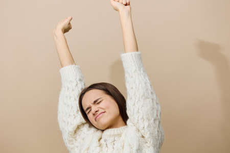 a tired woman stands and looks straight at a light brown background in a white sweater, stretches...